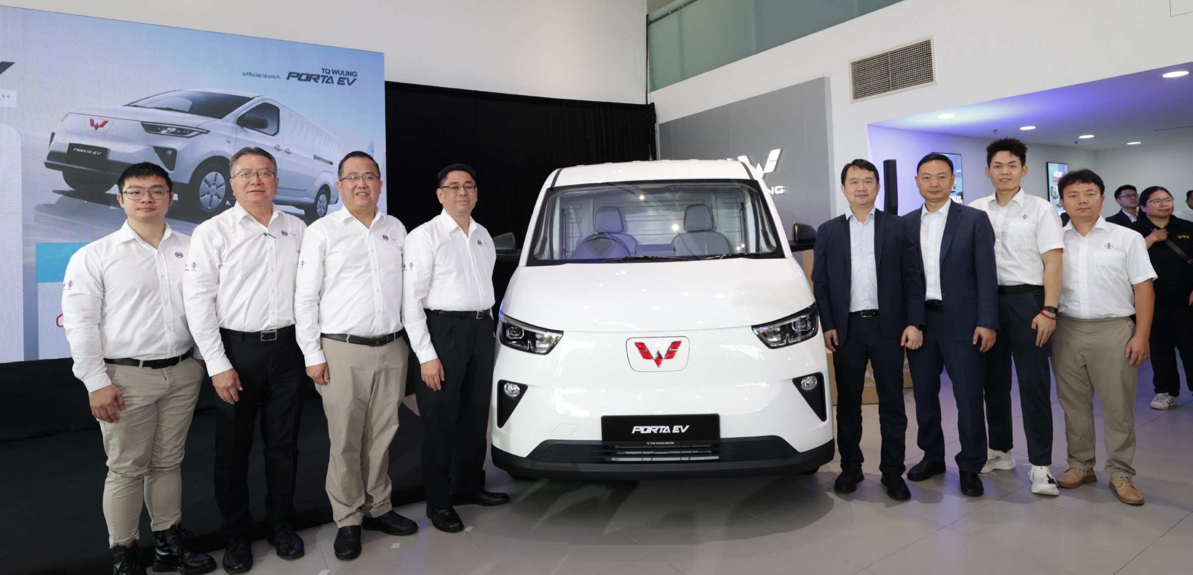 室内展厅里一辆白色 Porta EV 厢车正面朝镜头停着，车头中间是红色五菱车标，车牌位置写「PORTA EV」；车身两侧都站着人，左边四人穿白衬衫，右边两人穿深色西装、两人穿白衬衫，后方另有几个人；左后方立着一张展板，印着「official launch TQ WULING PORTA EV」和同一款白色厢车